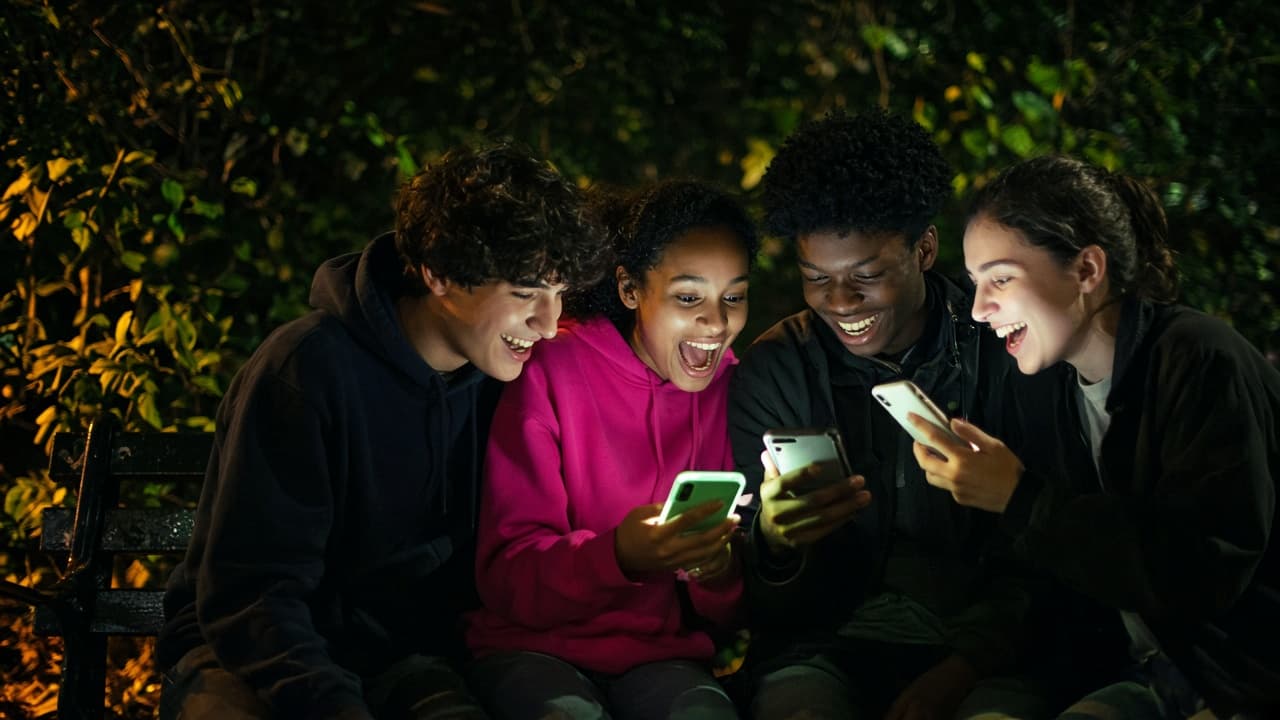 Friends laughing at their phones on a bench at night